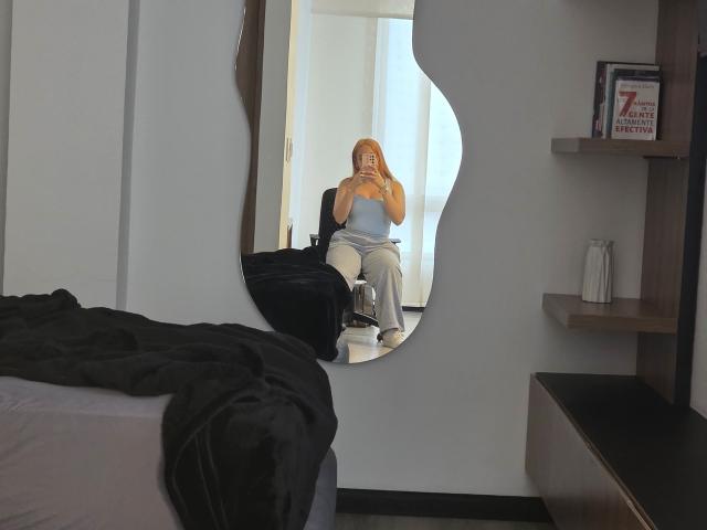 AlicePrice with orange hair, small tits, and ass, sitting in a chair, reflected in a wavy mirror, wearing a blue top and white pants, holding a phone.