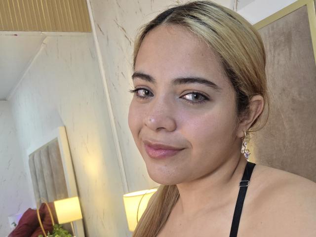 AlicePrice has blonde hair, brown eyes, and a slight smile. She's wearing black earrings and a strap. Her pinkish lips are soft and she looks cute.