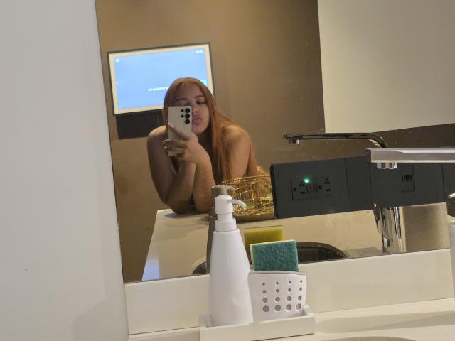 AlicePrice, with long red hair, takes a mirror selfie in a bathroom. She's topless, showing her tits, leaning on a white sink with a soap dispenser in front.
