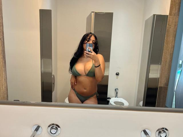 AlicePrice in a green bikini, showing her large tits and curvy ass, taking a mirror selfie in a bathroom with a phone. Her long black hair is down.
