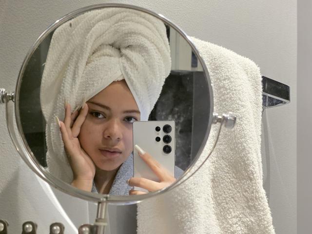 AlicePrice with a white towel wrapped around her head, holding a phone with three cameras, taking a mirror selfie, her fingers resting on her cheek, looking relaxed.