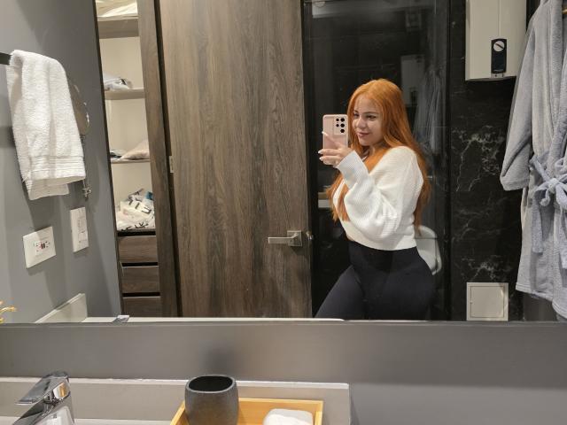 AlicePrice with long red hair, white sweater, and black pants, taking a mirror selfie in a bathroom, showing her curvy ass and cute smile. Her phone is pink.