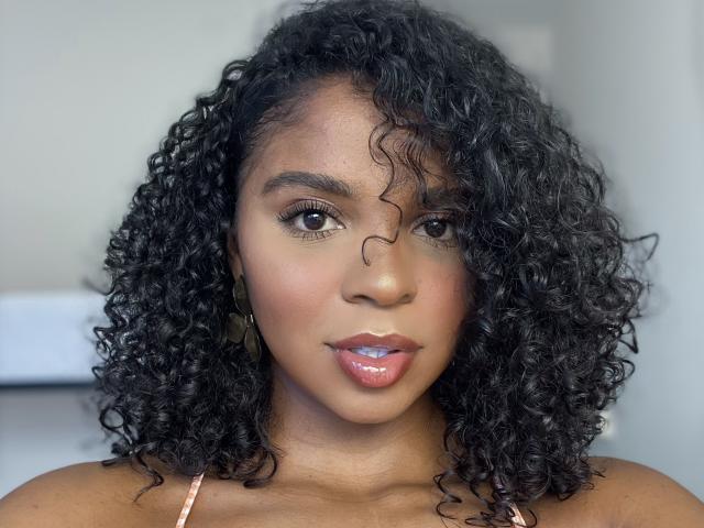 LisaMendez heeft donker krullend haar, bruine ogen en glanzende, roze lippen. Ze draagt een roze topje met spaghettibandjes, waardoor haar gladde, bruine huid en subtiele make-up zichtbaar zijn..
