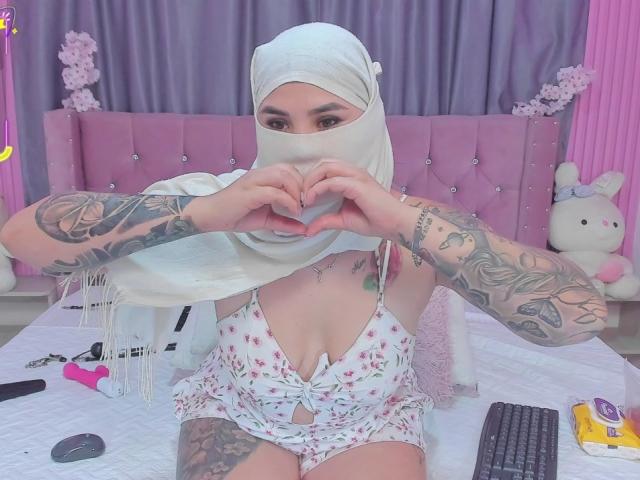 MalakCote with white hijab, floral lingerie, tattooed arms, heart gesture, pink bedroom, pink bed, white stuffed bunny, vibrator, remote, keyboard, and yellow packaging on bed.