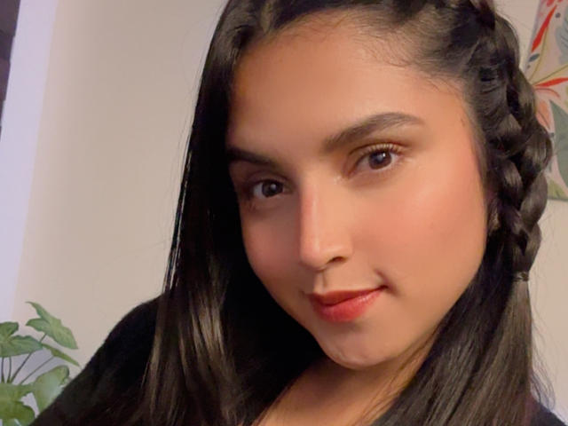 VeronicaFire69 has a cute face with dark brown eyes, red lipstick, and long black hair in braids. She's smiling softly with a slightly chubby nose.
