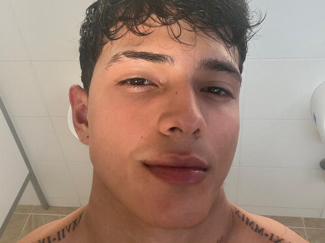 LyannG has a cute, slightly sweaty face with curly black hair, brown eyes, and a subtle smile. Tattoos are visible on his shoulders. He's shirtless in a bathroom.