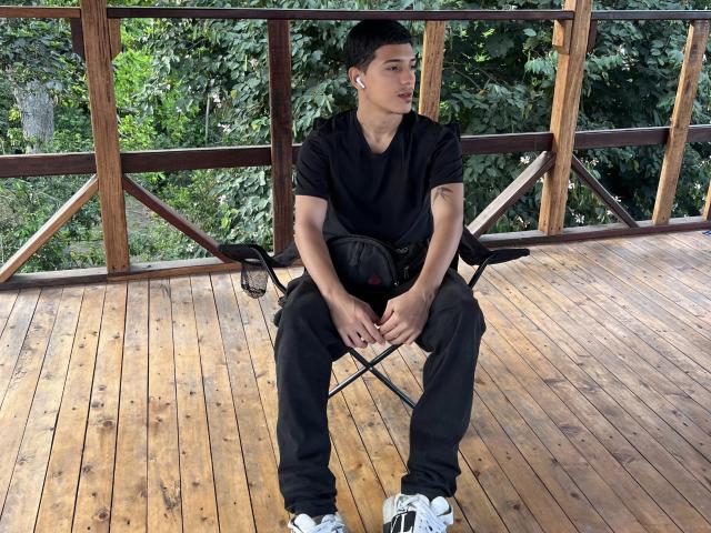 LyannG sitting on a wooden deck, legs apart, hands on black backpack, wearing black shirt, pants, white sneakers, wooden railing, green leafy background, short black hair, tattoo on right arm.