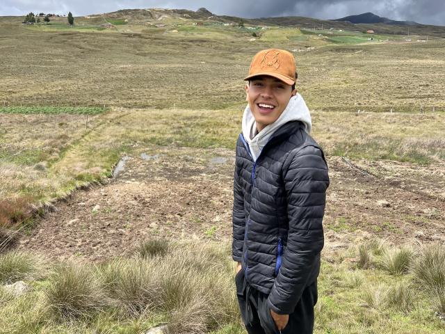 LyannG in a brown cap, black puffy jacket, and white hoodie, smiling with hands in black pants, standing in a grassy, rocky landscape with cloudy skies.