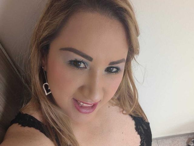 AlinaHill with light brown hair, blue eye makeup, and black lace top, smiling at the camera, showing her pink lips and wearing heart-shaped earrings.