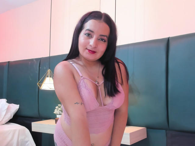 AnnaLeah, con cabello negro largo, un sujetador de encaje rosa y ropa interior a juego, sonriendo a la cámara, mostrando sus pechos, su trasero y un tatuaje pequeño en su brazo derecho..