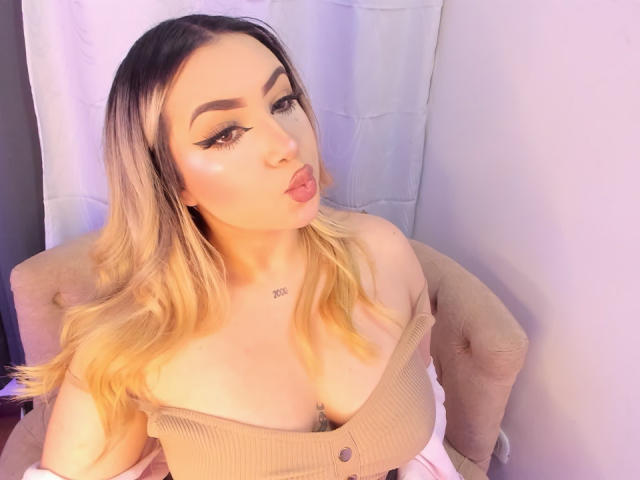 MelissaLust with blonde hair, pouty lips, and thick eyebrows, wearing a beige off-shoulder top, showing her large tits, sitting in a chair, with a white curtain behind.