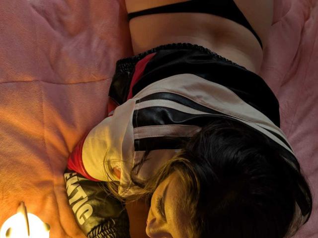KimyR lying on pink bed, black panties, black and white satin top, red accents, brown hair, small butt, visible black thong, soft light, pillow with green pattern.