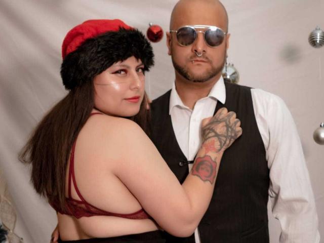 SektethLilith with big tits in red lace bra, Santa hat, and tattooed arm, stands next to bald dude in vest, white shirt, and sunglasses. He's looking serious.