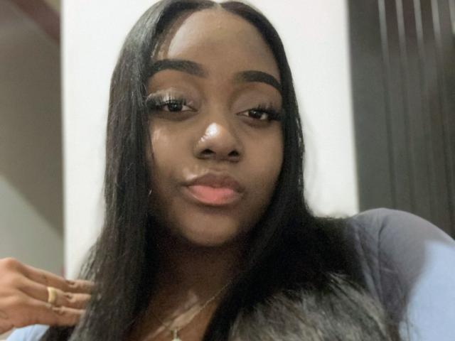 EbonnyMichell has long black hair, dark skin, and full pink lips. She's wearing a blue shirt, has a gold necklace, and is looking slightly to the side with a subtle smile.