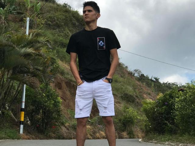 KevynStone stands with hands in white shorts, wearing a black T-shirt, a red phone icon on chest, and a black watch, against a green, hilly, cloudy background.