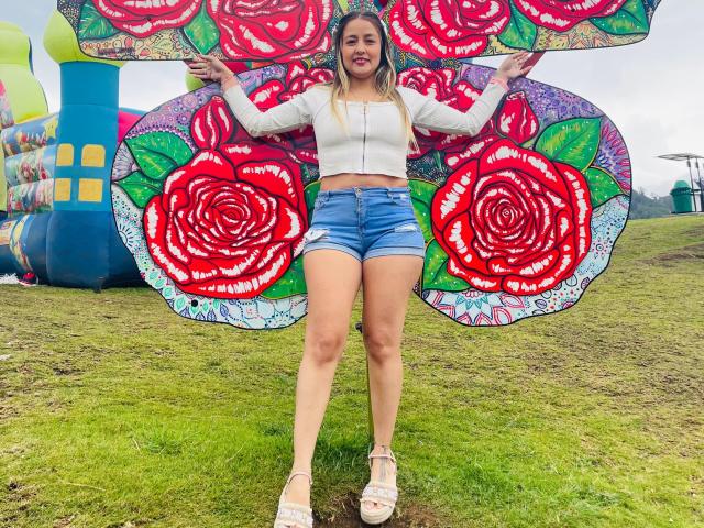 SaritaCaramelo with blonde hair, white crop top, blue shorts, white heels, standing on grass, arms out, holding colorful rose-patterned fabric, smiling, outdoors, grassy field, overcast.