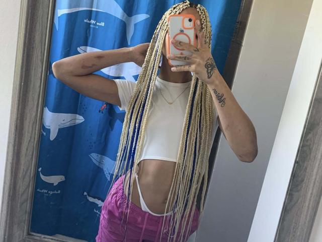 DolldSlov with long blonde braids, white crop top showing a sliver of their tits, pink pants, and tattoos, taking a mirror selfie with an orange phone.