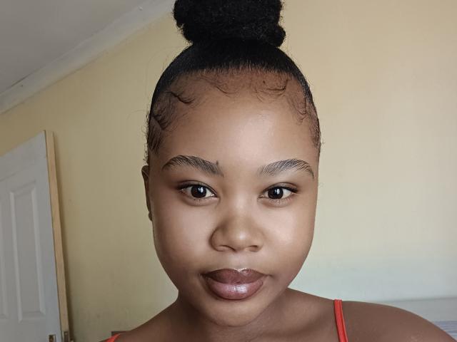 Brielly has dark brown skin, curly black hair in a bun, thick eyebrows, glossy brown lips, and is wearing a red spaghetti strap top.