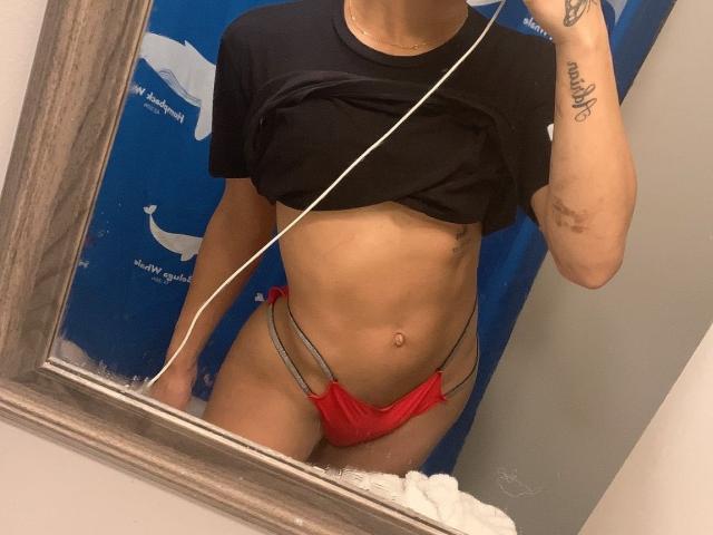 DolldSlov in a mirror selfie, wearing a black crop top, red string bikini bottom, showing a flat, toned stomach, and a tattoo on their right forearm.