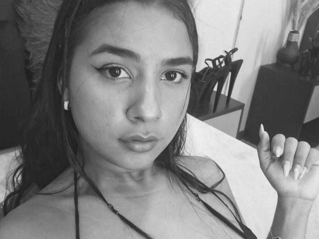 Black and white photo of MariamLara with long dark hair, wearing a halter top, showing her face and raised hand with manicured nails, wearing small white earrings.