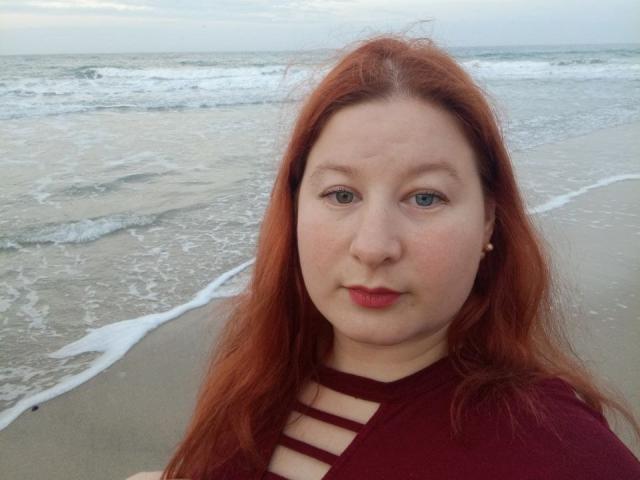 LizaLina has red hair, blue eyes, and pale skin with a slight blush. She's wearing a maroon top with strap details. Her red lipstick matches her hair.