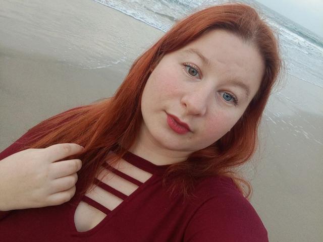LizaLina with red hair, fair skin, and green eyes, wearing a maroon top with strappy cutout, holding her hair, standing on a beach with gentle waves.