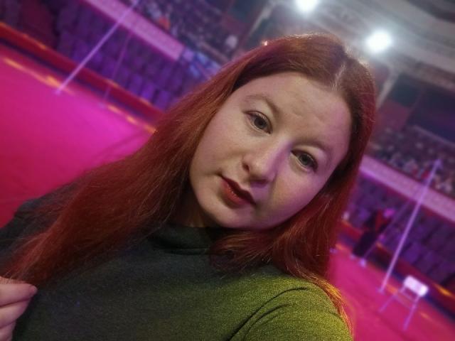 LizaLina has red hair, green eyes, and freckles, wearing a green shirt, with a slight smile, in a brightly lit indoor arena with pink and purple lighting.