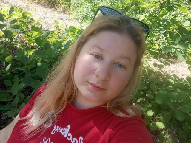 LizaLina with pale skin and blonde hair, wearing a red shirt, has a soft smile and is standing in green foliage, with black sunglasses on her head.