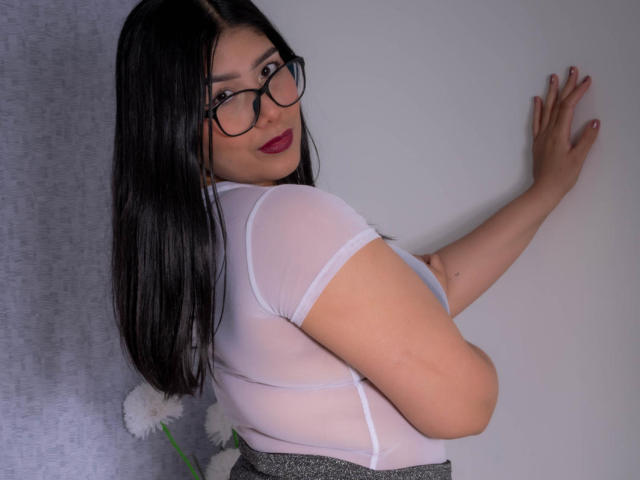 AlexaKiim with long black hair, glasses, and red lipstick, wearing a sheer white top, gray skirt, and touching a wall, showing her ass. White flowers in the background.
