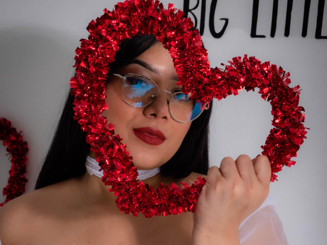 AlexaKiim with red heart-shaped tinsel, glasses reflecting blue, red lipstick, white choker, holding tinsel, black hair, fair skin, smiling, topless, showing bare shoulders.