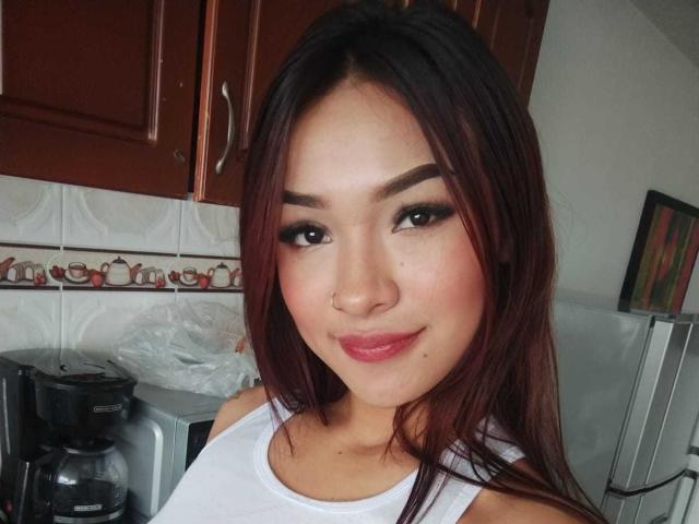 SashaHans has medium brown skin, red-brown hair, black eyeliner, and pink lipstick. She's wearing a white tank top, showing her small tits. She's smiling softly.