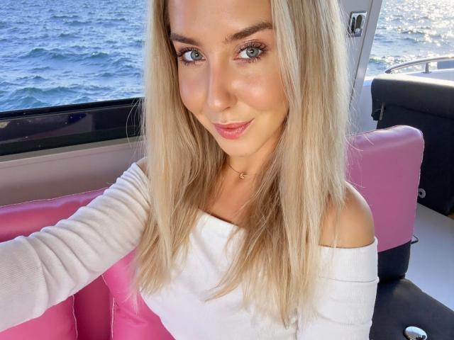Kia-rey-hot with blonde hair, blue eyes, and a white off-shoulder top, smiling on a boat with pink seats, showing her tits slightly. Ocean in background.