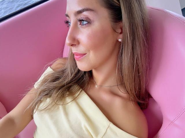 Kia-rey-hot with blonde hair, pink lipstick, and pearl earrings, sitting in a pink chair, wearing a yellow off-shoulder top, smiling softly, looking to the side.