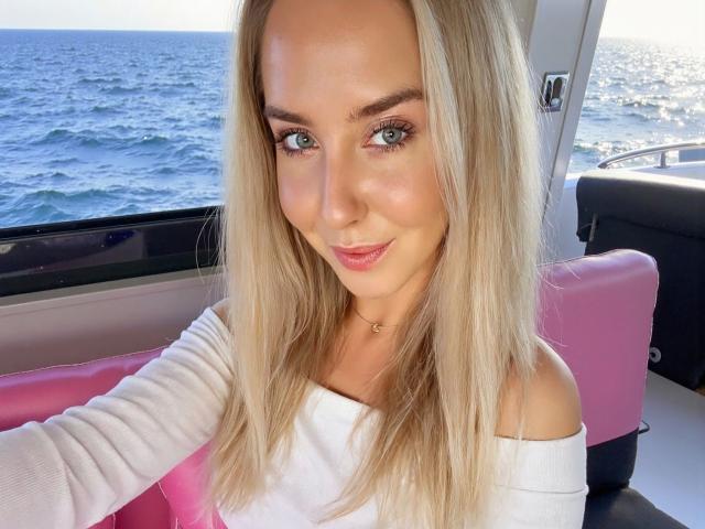 Kia-rey-hot has blonde hair, blue eyes, and is smiling with pink lips. She's wearing a white off-shoulder top, sitting on a pink seat by the ocean.