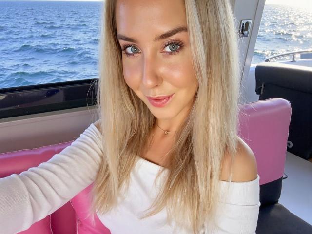 Kia-rey-hot with blonde hair, blue eyes, and a cute smile, wearing a white off-shoulder top, sitting on a pink seat on a boat, with the ocean in the background.