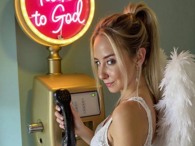 Kia-rey-hot with angel wings, blonde ponytail, white lace dress, holding black phone, smiling at camera, neon sign "to God" in background, green wall, golden machine.