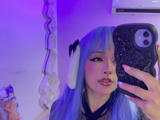 CarlaMiceli with blue hair, white bunny ears, and pink lipstick, holding a glittery phone, winking, purple-lit room, visible air conditioner, cute stuffed toy on left.
