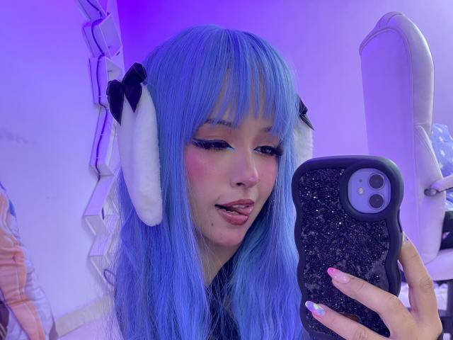 CarlaMiceli with blue hair, white headphones, and pink blush, holding a glittery phone, sticking out her tongue. Purple lighting. Her nails are multicolored. She's taking a selfie.