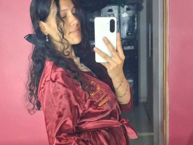A selfie of SoftCuteGirl with long, wavy black hair and a confident smile. She holds a white smartphone in her right hand, partially covering her face. She wears a shiny, deep red satin robe with embroidered text on the left chest. Her left wrist has a thin, black tattoo line. The pink wall in the background contrasts with the dark hair and red robe. The composition focuses on her upper body, capturing the reflection in a mirror.