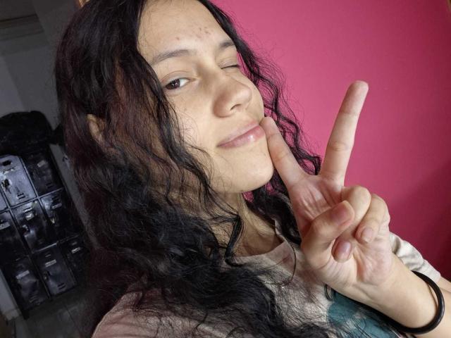 SoftCuteGirl with long black wavy hair, winking, making a peace sign with her right index finger, wearing a white shirt, pink background, slight acne on her cheek.