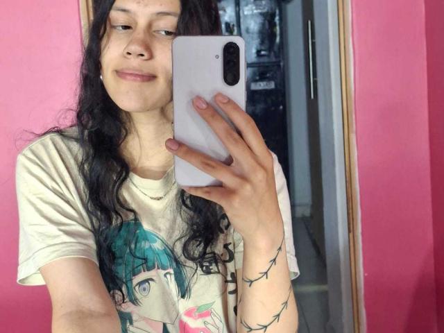 SoftCuteGirl with long black hair, wearing a white tee with a green-haired anime girl, holding a white phone, smiling, pink wall in the background, tattoo on right arm.
