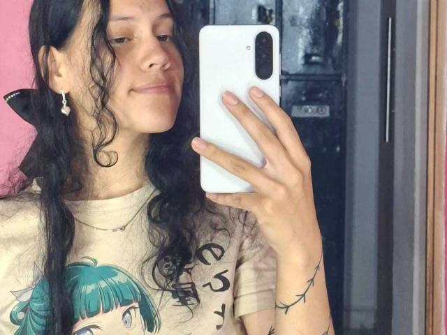 SoftCuteGirl with wavy black hair, small earrings, and a beige t-shirt with a green-haired graphic, holding a white phone, smiling slightly, reflecting in a mirror.