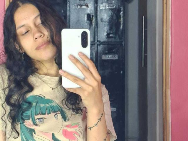Selfie of SoftCuteGirl with long, curly black hair and light brown skin. She wears a beige t-shirt with a green-haired anime girl graphic. She holds a white iPhone in her right hand, slightly smiling. Background shows a dark, metallic locker. SoftCuteGirl has a slender build and wears a thin, pink garment on her left arm. The pink wall on the right adds contrast.
