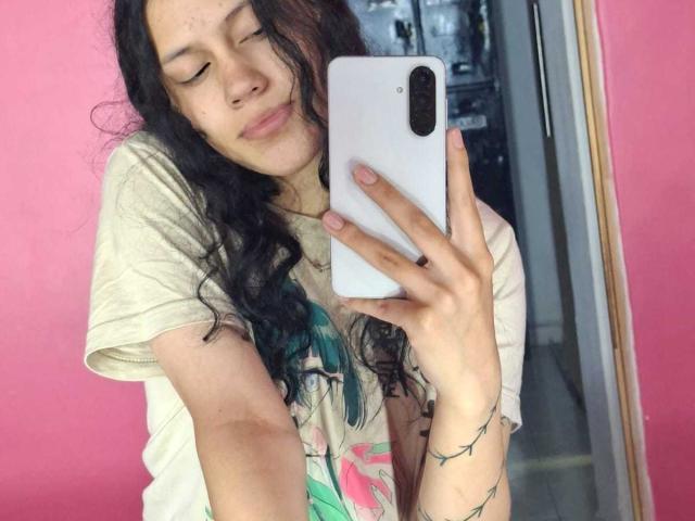 SoftCuteGirl with wavy black hair, light skin, and a small nose piercing, taking a selfie with a white phone, wearing a beige shirt with green and pink patterns.