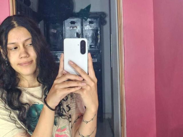 SoftCuteGirl with long, wavy black hair, wearing a beige graphic tee, taking a selfie with a white phone, smiling slightly, in a pink-walled room, showing a small scar.