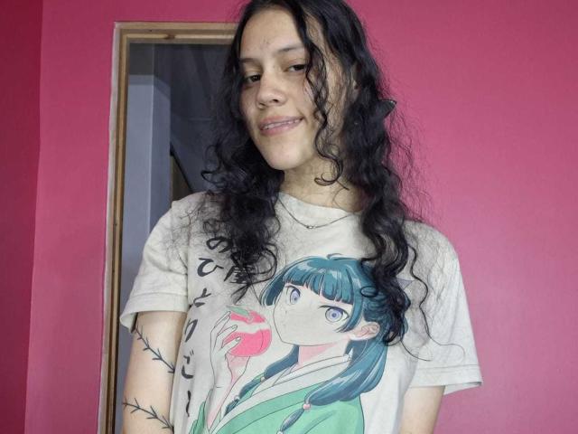 SoftCuteGirl with curly black hair, light skin, small tits, wearing a white anime print tee, holding a pink apple, standing against a pink wall, smiling gently.