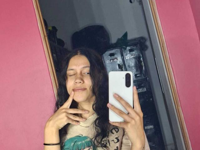 SoftCuteGirl with long curly brown hair, eyes closed, smirking, finger on chin, holding a white phone, pink wall, mirror selfie, wearing a beige shirt with a green graphic.