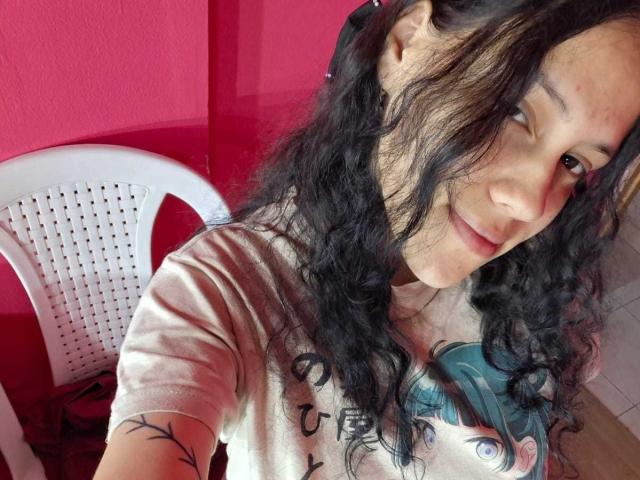 SoftCuteGirl with curly black hair, light brown skin, and a small tattoo on her arm, wearing a beige t-shirt with anime print, smiling at the camera. Red wall.