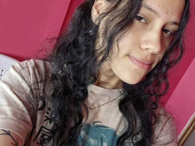 SoftCuteGirl with long, wavy black hair, light brown skin, small nose, subtle smile, wearing a beige t-shirt, visible tattoo on right arm, pink wall in background.
