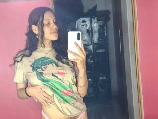 SoftCuteGirl with curly dark hair, wearing a tee with anime girl print, holding phone, pink walls, mirror reflection, pink panties, small tattoos, confident smile, gentle hand on belly.